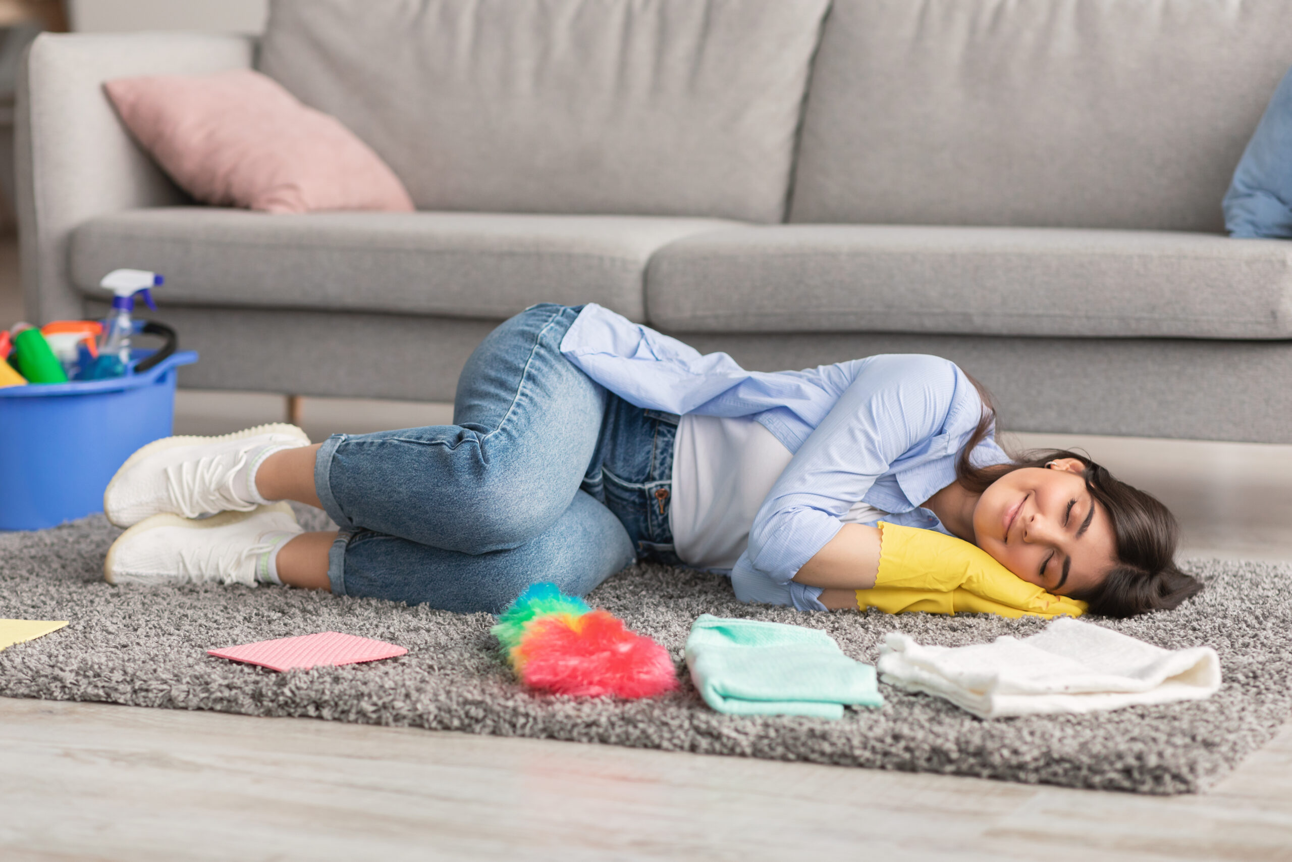 Tired millennial housewife sleeping on gray rug carpet in living room, surrounded by cleaning supplies, copy space. Overworked young lady napping, exhausted after domestic duties, needing rest